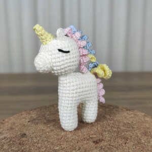 May include: A white crocheted unicorn with a yellow, pink, and blue mane. The unicorn has a closed eye and a small horn.
