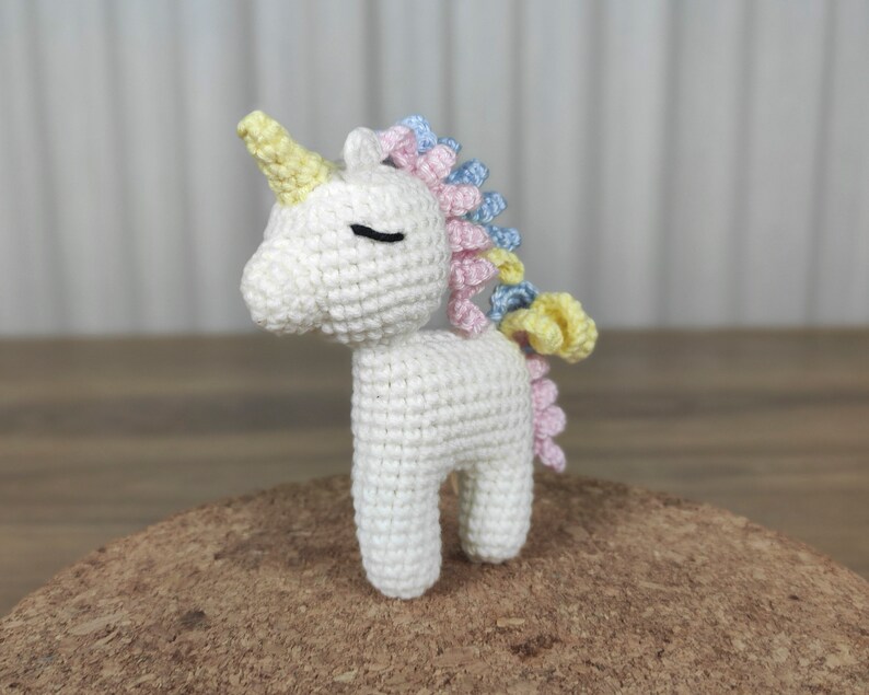 May include: A white crocheted unicorn with a yellow, pink, and blue mane. The unicorn has a closed eye and a small horn.
