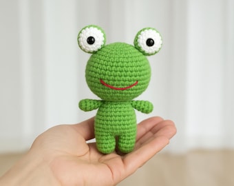 Op de afbeelding: Een kleine groene gehaakte kikker met witte ogen en een rode glimlach. De kikker zit in de palm van een hand.