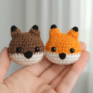 Puede incluir: Dos pequeños peluches de zorro de crochet, uno marrón y otro naranja, con caras blancas y ojos negros. Los peluches están sostenidos en la mano de una persona.