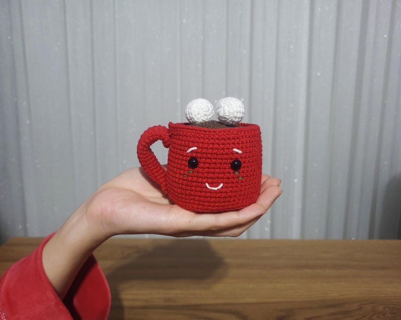 Op de afbeelding: Een handgemaakte rode gehaakte mok met een lachend gezicht, zwarte kraaloogjes en twee witte pompons bovenop. De mok wordt in een hand gehouden, wat het grillige ontwerp benadrukt. Een charmant, handgemaakt item.