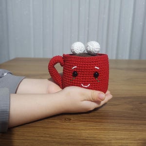 Op de afbeelding: Een handgemaakte, rode gehaakte mok met een lachend gezicht en twee witte, pluizige ballen bovenop. De mok heeft een oor en is gevuld met bruin materiaal, mogelijk koffie of warme chocolademelk. De mok wordt in de handen van een persoon gehouden.