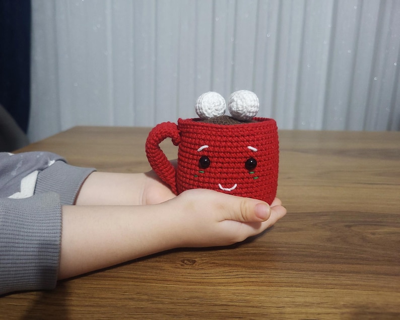 Op de afbeelding: Een handgemaakte, rode gehaakte mok met een lachend gezicht en twee witte, pluizige ballen bovenop. De mok heeft een oor en is gevuld met bruin materiaal, mogelijk koffie of warme chocolademelk. De mok wordt in de handen van een persoon gehouden.