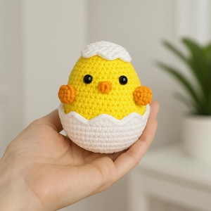 Op de afbeelding: Een gehaakt kuiken dat uit een witte eierschaal kijkt. Het kuiken is geel met oranje pootjes en zwarte ogen.