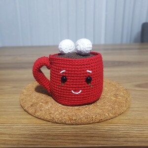 Op de afbeelding: Een handgemaakte rode gehaakte mok met een lachend gezicht, zwarte ogen en twee witte marshmallow-accenten. De mok staat op een kurken onderzetter, wat een gezellig, handgemaakt thema suggereert.