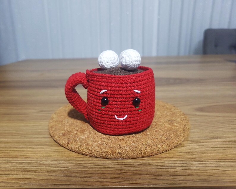 Op de afbeelding: Een handgemaakte rode gehaakte mok met een lachend gezicht, zwarte ogen en twee witte marshmallow-accenten. De mok staat op een kurken onderzetter, wat een gezellig, handgemaakt thema suggereert.