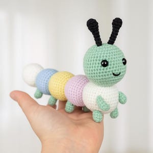 Può includere: Un giocattolo a forma di bruco fatto a crochet con una testa verde, antenne nere e un corpo bianco, azzurro, giallo e rosa.