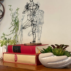 May include: A wire sculpture of a person in a yoga pose, on top of two books, with a succulent plant in a grey hand-shaped planter on a wooden shelf.