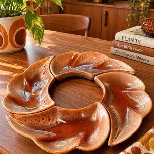 May include: A set of four curved, crescent-shaped ceramic dishes arranged in a circular formation. Each dish features a glossy, variegated glaze transitioning from a deep, rich burnt orange in the center to a lighter, speckled tan at the edges.