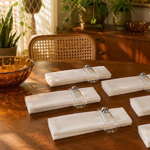 May include: A set of six rectangular white cloth napkins, each secured with a clear acrylic napkin ring featuring a polished silver-tone metal ball accent. The napkins are neatly folded and arranged on a wooden surface, showcasing a clean design.