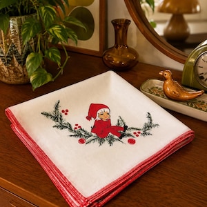 Peut inclure: Une serviette en tissu carrée blanche avec une bordure cousue rouge, ornée d'un motif central représentant un petit personnage en tenue et bonnet rouges assis sur une branche de pin verte avec des baies rouges, disposé de manière symétrique.