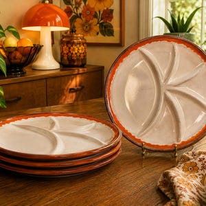 Peut inclure: Un ensemble d'assiettes rondes en céramique avec un centre crème divisé en cinq sections distinctes par des crêtes surélevées. Chaque assiette présente un large bord orange texturé. Une assiette est posée verticalement sur un support.