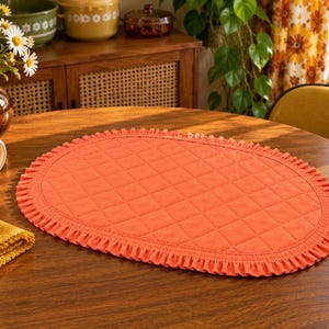 May include: An oval-shaped orange placemat featuring a quilted diamond pattern across the centre and a ruffled fabric border along the entire edge. The placemat is constructed from a solid-coloured fabric and sits flat on a wooden surface.