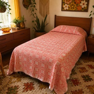 May include: A twin-sized bed featuring a coral pink and white woven bedspread with a repeating geometric square pattern. The bedspread covers the mattress and hangs over the sides, paired with a matching pillow sham covering the head of the bed.