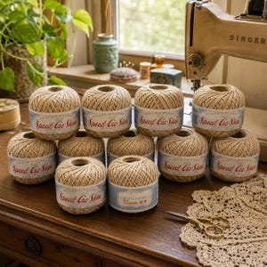 Peut inclure: Onze pelotes de fil à crocheter en coton beige Coats & Clark Speed-Cro-Sheen disposées sur une surface en bois. Chaque pelote porte une étiquette bleu clair avec le texte rouge Speed-Cro-Sheen. Des ciseaux dorés reposent sur un napperon en dentelle.