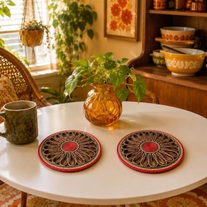 May include: Two round, woven coasters featuring a dark brown, petal-like pattern radiating from a solid red centre, finished with a thin red border. The coasters are placed on a white table next to a green ceramic mug and an amber glass vase.