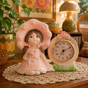 May include: A ceramic figurine of a girl wearing a large pink hat and a pink dress, holding a bouquet of flowers, positioned next to a round analog alarm clock with a pink frame, floral accents, and a green base labeled with the brand Equity.