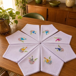 Peut inclure: Un ensemble de huit serviettes en tissu violet clair disposées en cercle. Chaque serviette présente un motif de paon brodé unique dans des couleurs vives, notamment bleu, rose, jaune et gris, perché sur une petite branche verte.