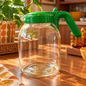Puede incluir: Una jarra de cristal transparente con una tapa de plástico verde brillante y un asa a juego del mismo color. La jarra tiene un cuerpo cilíndrico ancho con un acabado liso y transparente, diseñada para verter líquidos con un asa lateral firme.