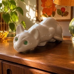 May include: A white ceramic figurine of a crouching cat with smooth, glossy surfaces and distinct, bright green eyes. The feline sculpture is depicted in a low, prowling posture with its head extended forward and ears alert, showcasing a minimalist form.