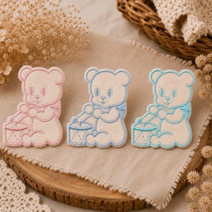 Peut inclure: Trois écussons brodés représentant un ours en peluche assis jouant du tambour. L'écusson de gauche est bordé de rose, tandis que ceux du milieu et de droite sont bordés de bleu. Chaque ours a un corps crème avec des détails faciaux cousus.