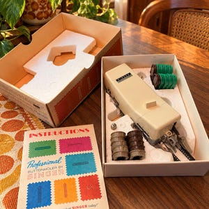 May include: A vintage Singer Professional buttonholer attachment set in a cardboard box with white polystyrene inserts. The kit includes a beige metal buttonholer, several plastic cams in green, brown, and grey, and an instruction booklet.