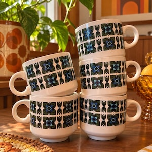 Puede incluir: Un conjunto de cuatro tazas de cerámica blanca apiladas en dos columnas. Cada taza presenta un patrón geométrico repetitivo en tonos verde oscuro, verde claro y azul. Las tazas tienen un cuerpo cilíndrico, un asa circular y una base.