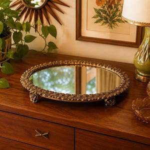 Puede incluir: Una bandeja redonda con superficie de espejo y un marco de metal dorado decorativo con un motivo de rosas detallado. La bandeja se apoya sobre tres pequeños pies ornamentados con forma de rosa, proporcionando una base elevada.