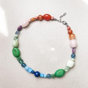 Könnte beinhalten: Eine farbenfrohe Perlenkette mit einer Vielzahl von Formen und Größen. Die Perlen sind in den Farbtönen Rot, Orange, Rosa, Lila, Grün, Blau und Weiß gehalten. Die Halskette hat einen silberfarbenen Verschluss.
