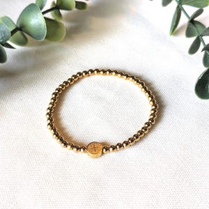 May include: A gold-toned beaded bracelet featuring a series of small, uniform metallic spheres strung together. The bracelet includes a single, slightly larger, flat circular charm positioned at the center, which displays a subtle engraved cross design.