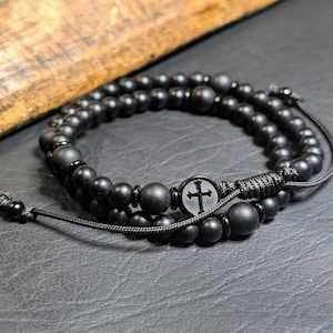 May include: A double-strand bracelet featuring matte black spherical beads of varying sizes, strung on a black cord. The design includes a circular charm with a black cross emblem and a sliding knot closure with small black beads at the ends.