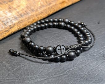 May include: A double-strand bracelet featuring matte black beads of varying sizes and a central circular charm displaying a black cross. The bracelet is constructed with a black cord and includes an adjustable sliding knot closure mechanism.
