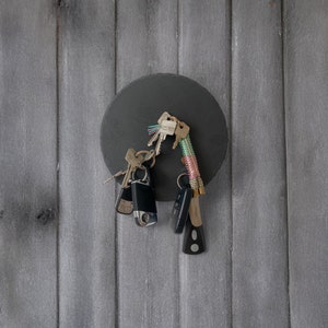 May include: A black round magnetic key holder with a variety of keys attached to it. The key holder is mounted on a gray wooden wall.