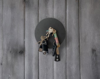 May include: A black round magnetic key holder with a variety of keys attached to it. The key holder is mounted on a gray wooden wall.