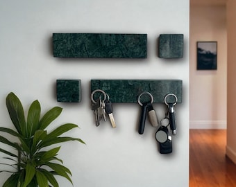 May include: A set of dark green marble key holders mounted on a white wall. The set includes two rectangular and two square pieces. Keys hang from the lower rectangular holder. A houseplant is visible in the lower left corner.