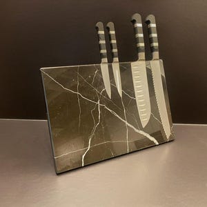 May include: A set of four stainless steel knives with striped handles displayed in a black marble knife block. The block has white veining. The knives include a paring knife, a utility knife, and a serrated bread knife.