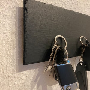 May include: A black slate key holder with a set of keys hanging on it. The keys include a car key fob with a Volkswagen logo.