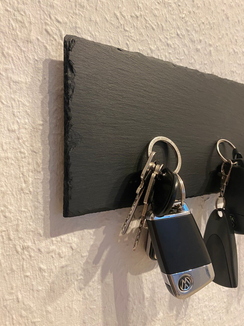 May include: A black slate key holder with a set of keys hanging on it. The keys include a car key fob with a Volkswagen logo.