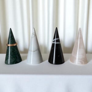 Pode incluir: Quatro suportes de anel em forma de cone. Os suportes são feitos de mármore verde, mármore branco, mármore preto e pedra rosa. Um anel de ouro repousa sobre o suporte de mármore verde.