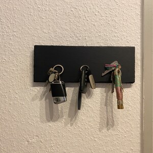 May include: A black key holder with three sets of keys hanging from it. The keys are attached to a variety of keychains, including a silver car key fob, a silver key ring with a small silver charm, and a colorful braided keychain.