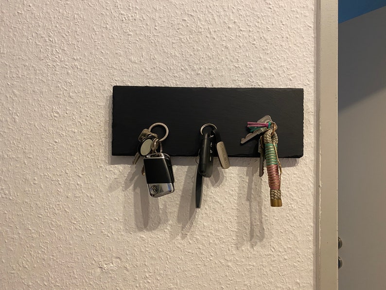 May include: A black key holder with three sets of keys hanging from it. The keys are attached to a variety of keychains, including a silver car key fob, a silver key ring with a small silver charm, and a colorful braided keychain.