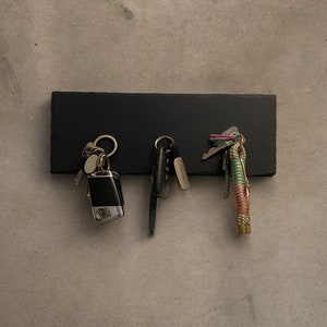 Op de afbeelding: Een zwarte leisteen sleutelhanger met drie sets sleutels die eraan hangen. De sleutels zijn een mix van zilver en goud, en één set heeft een kleurrijk touw.