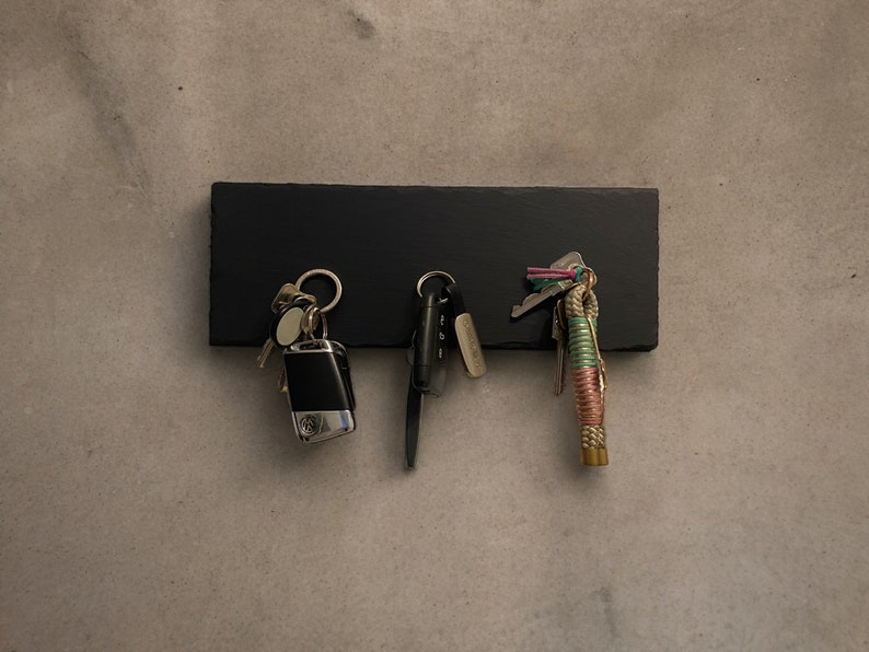 May include: A black slate key holder with three sets of keys hanging from it. The keys are a mix of silver and gold, and one set has a colorful rope attached.