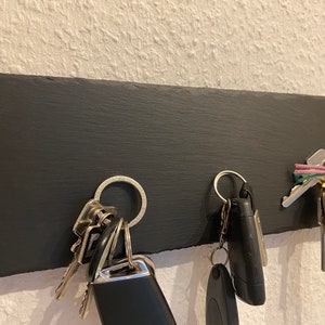 May include: A black slate key holder with a silver ring and keys hanging from it. There are two sets of keys, one with a black fob and the other with a silver fob. A pink and green lanyard with a gold charm is also hanging from the holder.