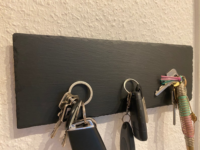 May include: A black slate key holder with a silver ring and keys hanging from it. There are two sets of keys, one with a black fob and the other with a silver fob. A pink and green lanyard with a gold charm is also hanging from the holder.