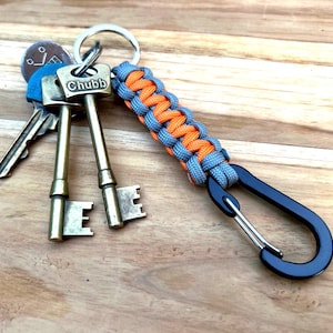 May include: A set of keys attached to a metal keyring and a braided orange and grey paracord keychain. The keys include one stamped with "Chubb". A black carabiner clip is also attached.