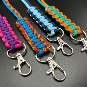 May include: Four colourful paracord lanyards with silver swivel snap hooks. The lanyards are in shades of pink, purple, blue, orange, and teal. The braided designs are visible, and the hooks are attached to the ends.