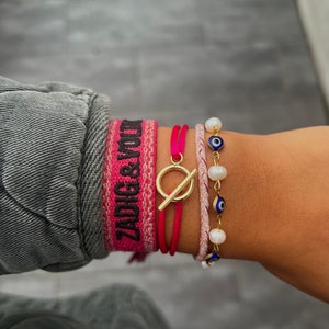 Peut inclure: Un ensemble de quatre bracelets, comprenant un bracelet tressé rose et blanc, un bracelet en cordon rouge avec un fermoir à glissière doré, un bracelet tressé rose et blanc avec un fermoir à glissière doré et un bracelet à breloques en perles blanches et en œil bleu.
