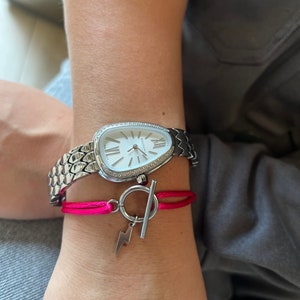 Peut inclure: Une montre-bracelet argentée avec un cadran blanc et des détails en diamants, associée à un bracelet rose vif orné d'un charm en forme d'éclair. La montre a un bracelet texturé. Le bracelet ajoute une touche de couleur vive.