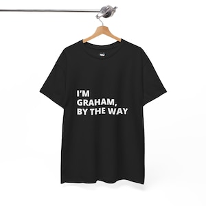 May include: Black t-shirt with the white text "I'M GRAHAM, BY THE WAY." The shirt is hanging on a wooden hanger suspended from a metal bar. The shirt has short sleeves and a crew neck.